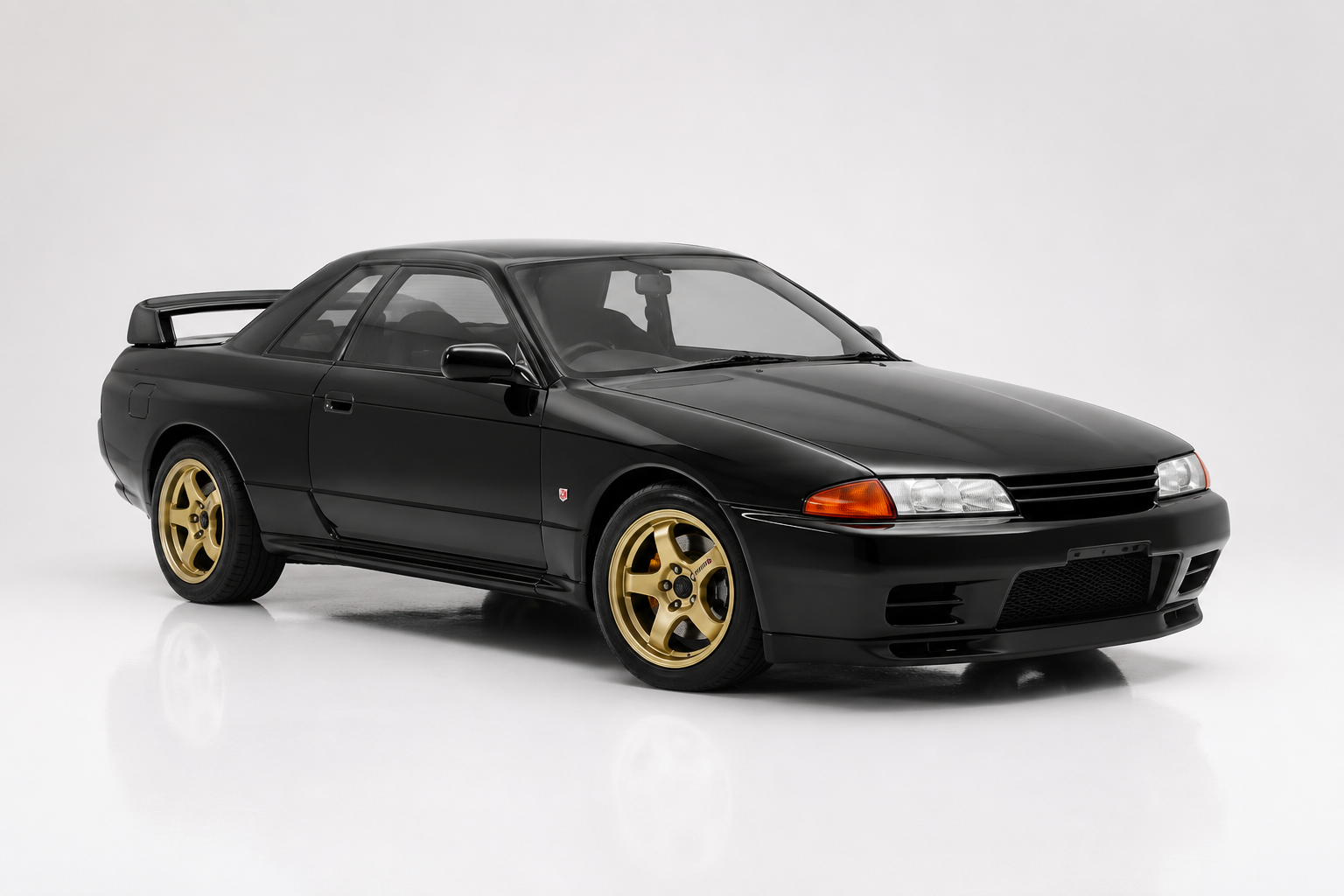Black Nissan Skyline GT-R R32 in a studio setting, front three-quarter view showing the boxy coupe body, twin round headlamps, small rear ducktail spoiler and gold multi-spoke wheels.