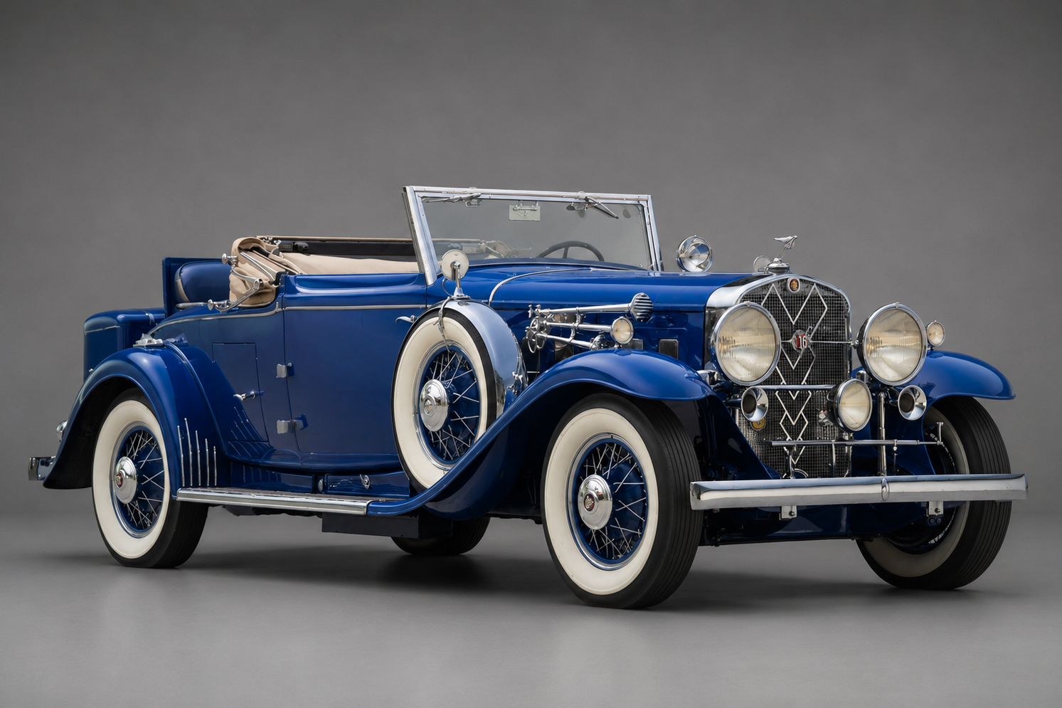 Deep blue Cadillac V-16 Series 452 convertible coupe with the top down, side-mounted spare wheel and whitewall tyres, front three-quarter studio view on a plain grey backdrop showing the V-16 crest grille, chrome twin headlamps and running boards.