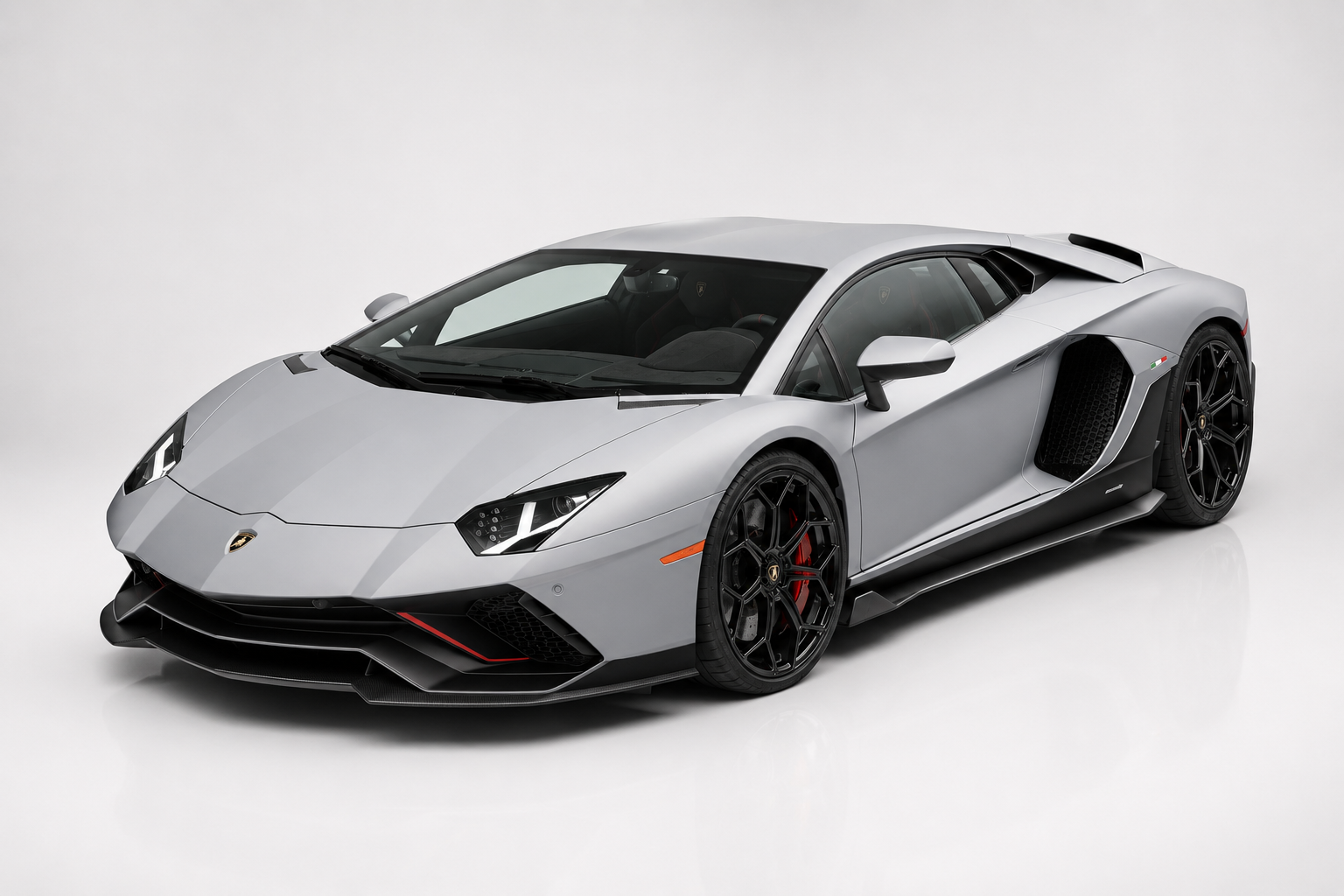 Silver-grey Lamborghini Aventador coupe in a bright studio, front three-quarter view showing the angular V12-era bodywork, Y-shaped LED headlamps, aggressive front splitter with red accents, side air intake with black mesh, Italian tricolour fender flash and black multi-spoke alloy wheels with red brake calipers.