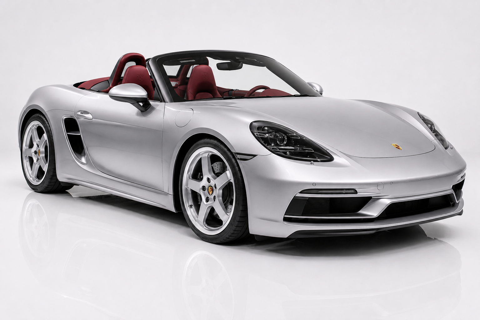 Silver Porsche 718 Boxster with red leather interior, roof down, front three-quarter view in a studio setting showing the low mid-engine roadster body, LED headlamps and multi-spoke alloy wheels.
