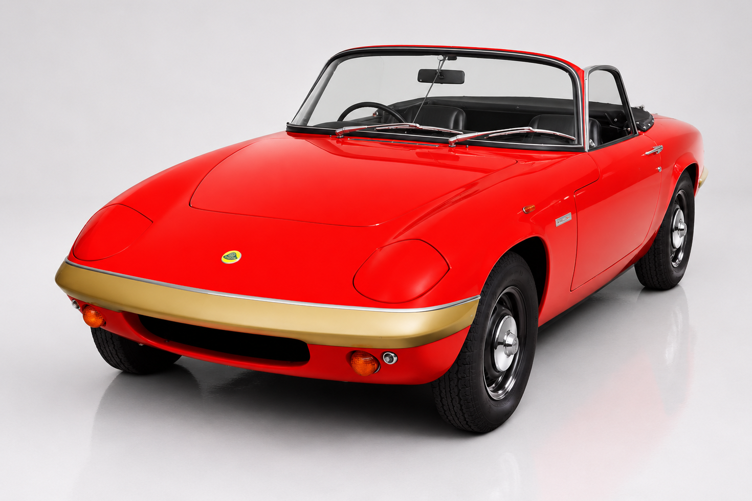Red Lotus Elan drophead coupé in a bright studio, front three-quarter view with the roof down showing the low pointed nose, pop-up headlights, painted gold nose band trimmed in chrome, small windscreen and black steel wheels — Colin Chapman's masterpiece and Lotus's first commercial success.