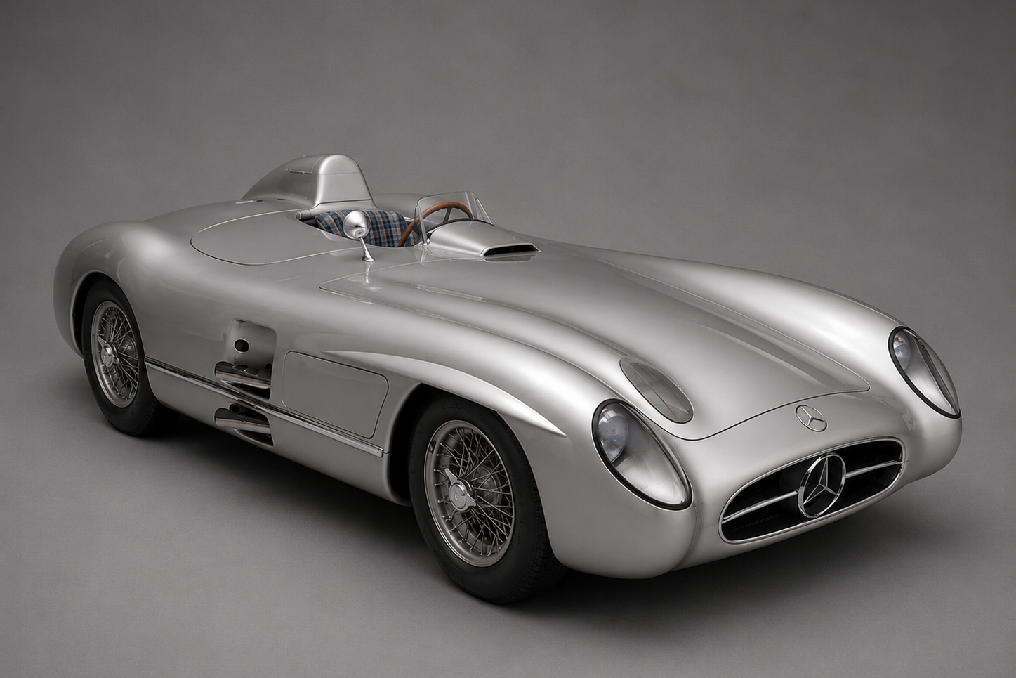 Studio three-quarter front view of a silver Mercedes-Benz 300 SLR open roadster with headrest fairing behind the driver, wire wheels, low-set Mercedes-Benz star grille and streamlined bodywork, on a plain grey backdrop.