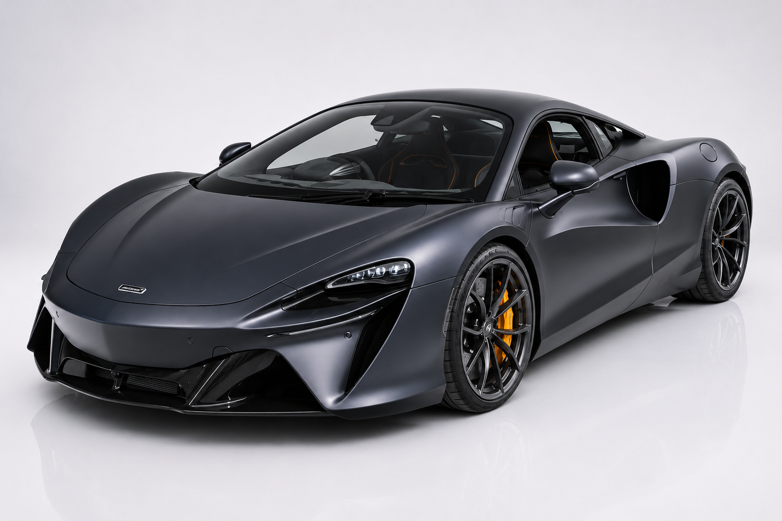 Dark matte-grey McLaren Artura in a studio setting, front three-quarter view showing the low mid-engine silhouette on the MCLA carbon platform, LED front lighting, prominent side air intake behind the door and dark multi-spoke wheels with yellow brake calipers — McLaren's first series-production V6 hybrid supercar.