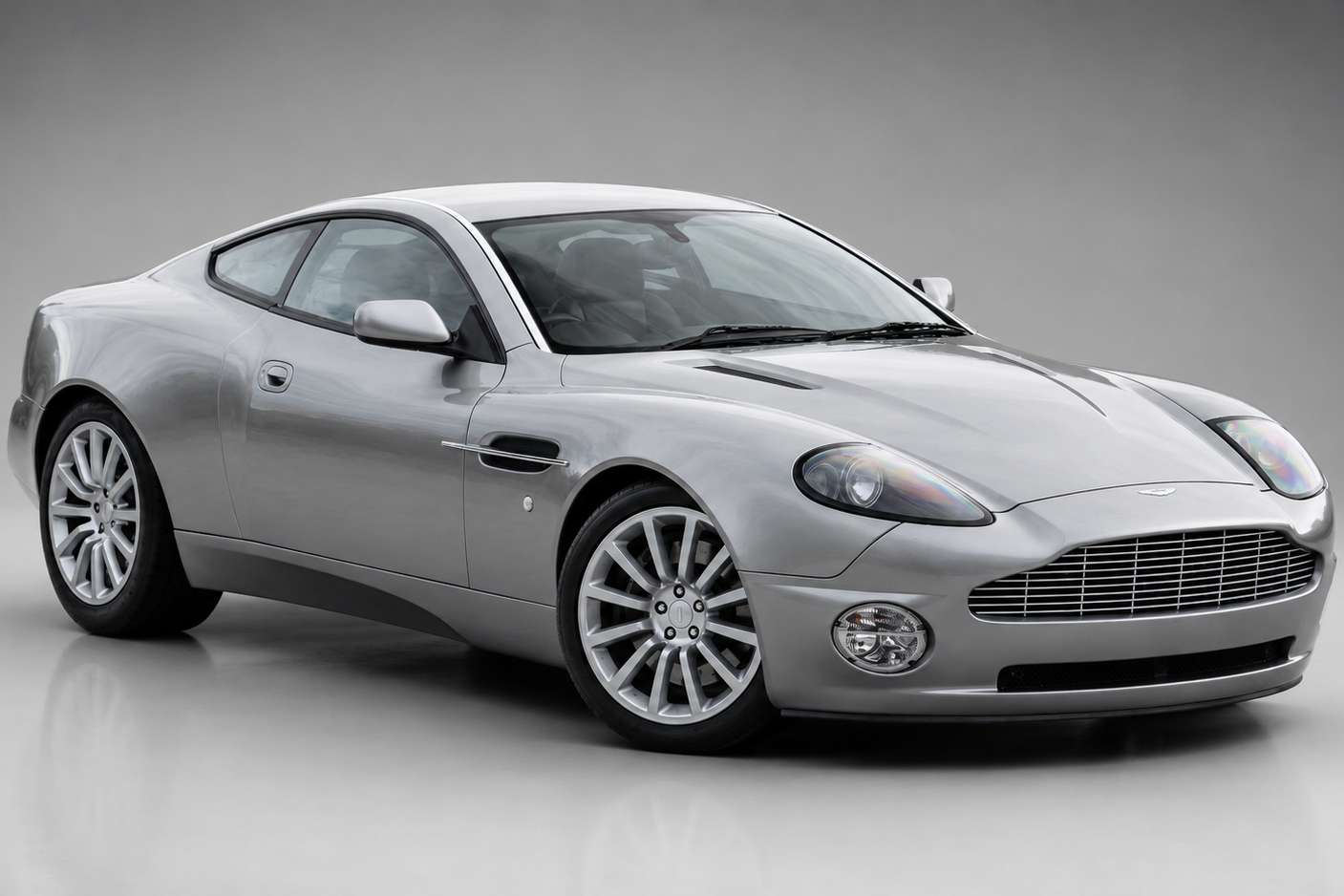 Silver Aston Martin Vanquish coupe in a grey studio setting, front three-quarter view showing the mesh grille, bonnet vents, side vent strake and multi-spoke alloy wheels.