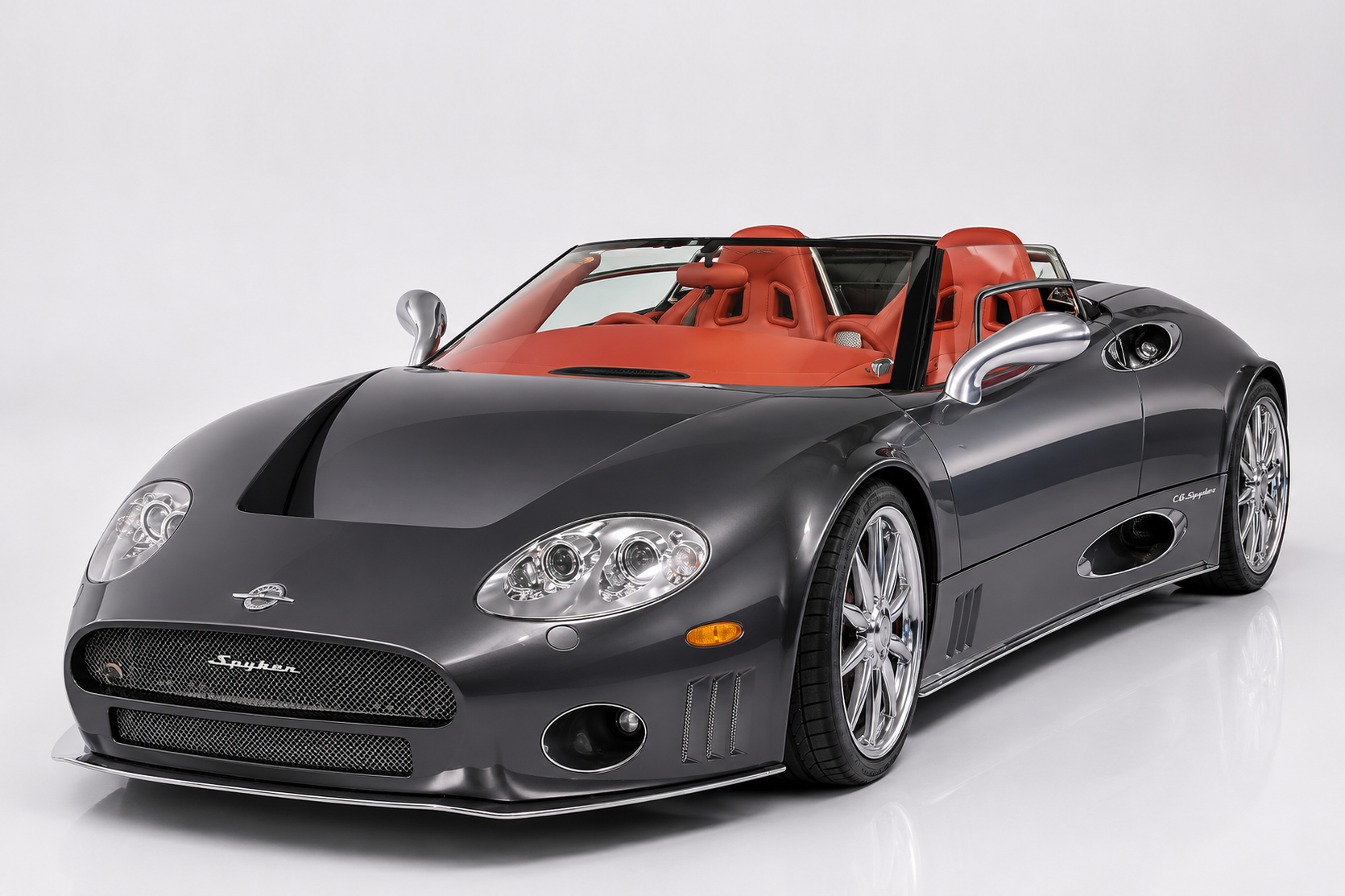 Dark grey Spyker C8 Spyder in a studio setting, front three-quarter view showing the low aluminium bodywork with exposed side exhausts, aviation-themed grille with Spyker script, quad round headlamps, red leather interior and multi-spoke chrome alloy wheels.