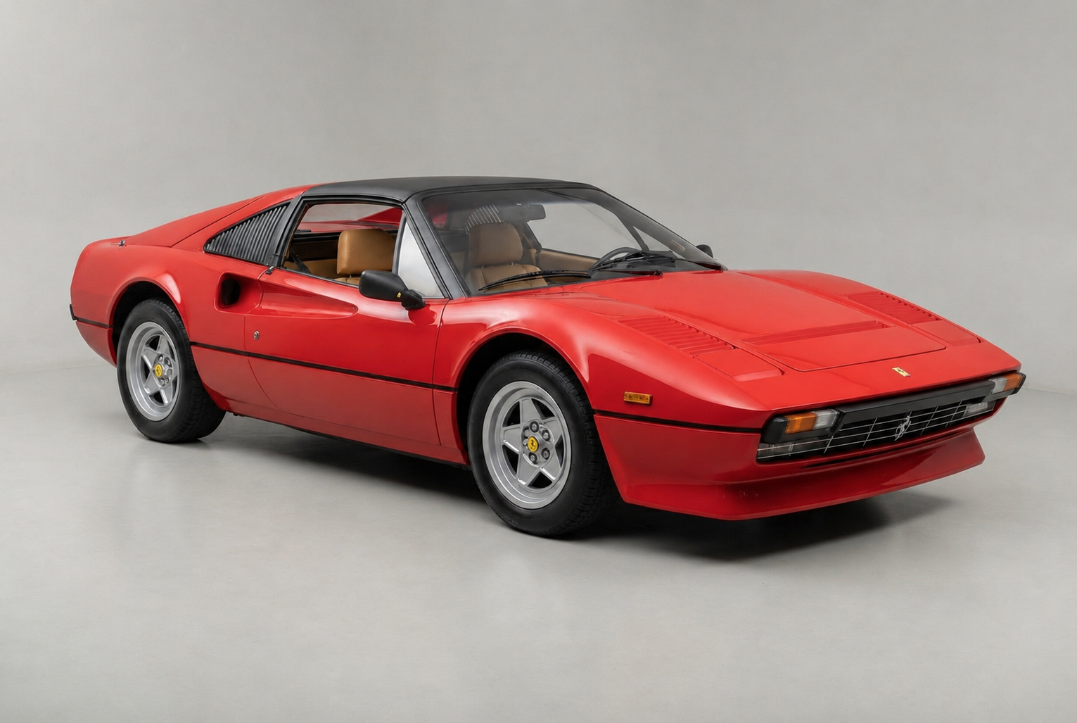 Red Ferrari 308 GTS, front three-quarter studio view on a pale grey backdrop, showing the Pininfarina mid-engined bodywork, the black removable targa roof panel, satin black louvred panels over the rear quarter windows, pop-up headlamps closed, five-spoke alloy wheels and tan interior.