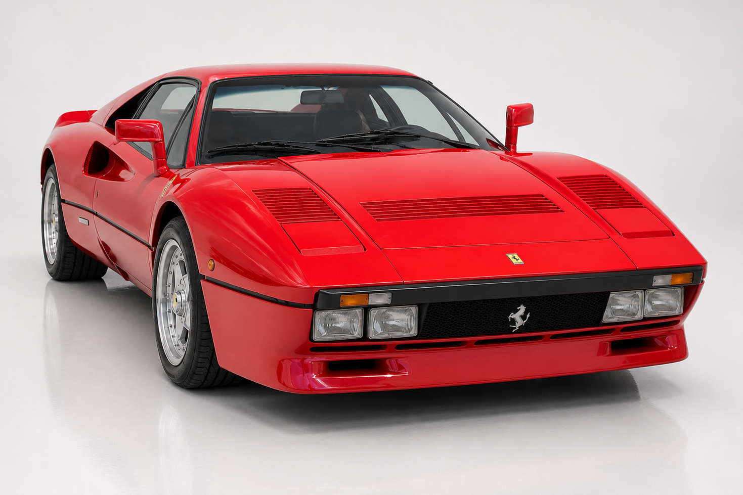 Red Ferrari 288 GTO coupe in a bright grey studio setting, front three-quarter view showing the four round headlamps set into the black front bumper band, black lower bumper with brake-cooling intakes, slatted bonnet vents, side NACA duct and five-spoke alloy wheels.