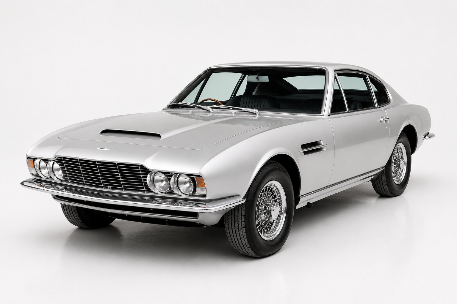 Silver Aston Martin DBS coupe in a grey studio setting, front three-quarter view showing the mesh grille flanked by quad round headlamps, chrome bumper, side vent strake and chrome wire wheels.