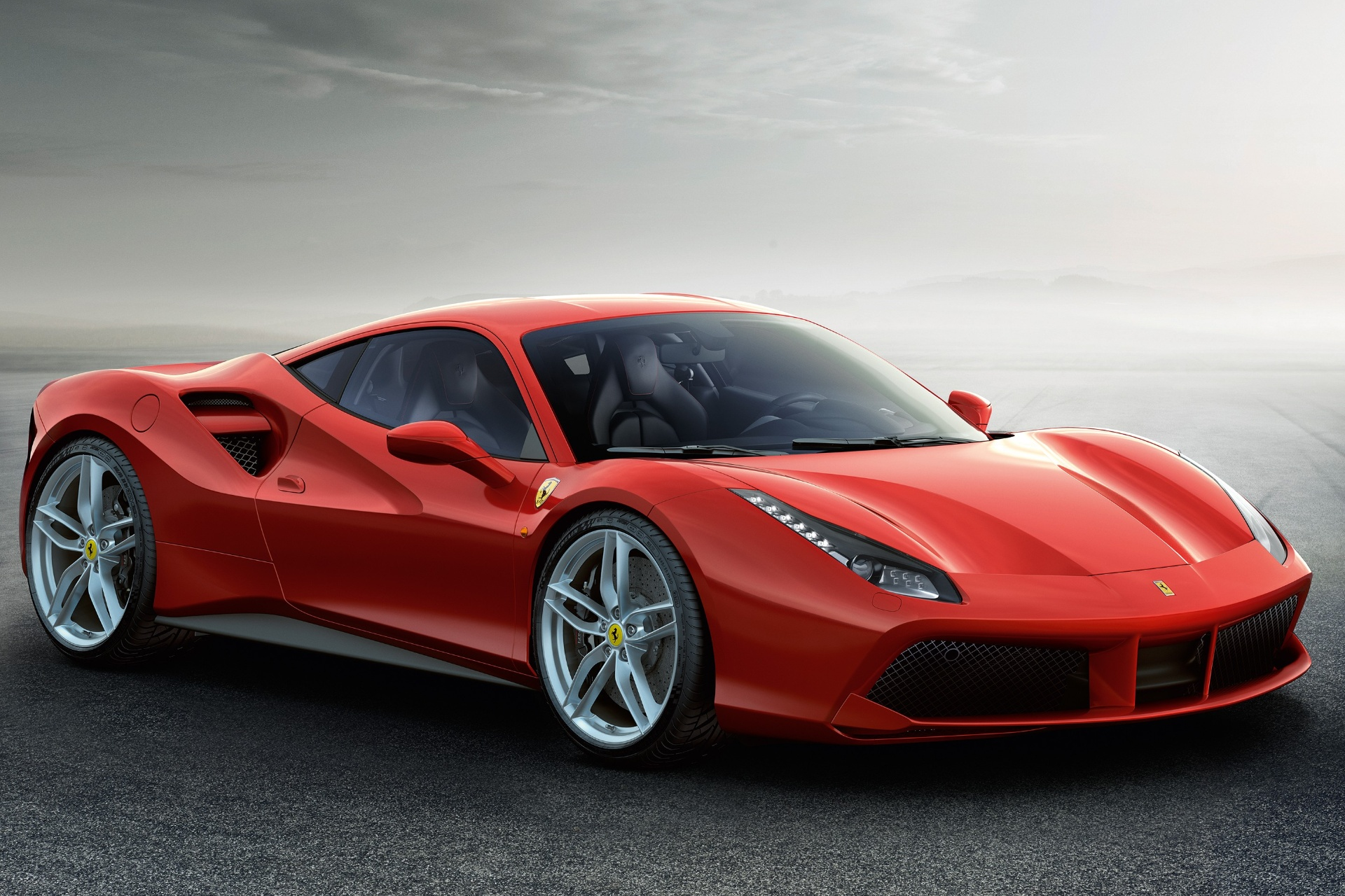 Red Ferrari 488 GTB, front three-quarter studio view on tarmac against a misty grey sky, showing the sculpted side intake with aero splitter, LED headlamps, silver five-spoke wheels and low mid-engined proportions.