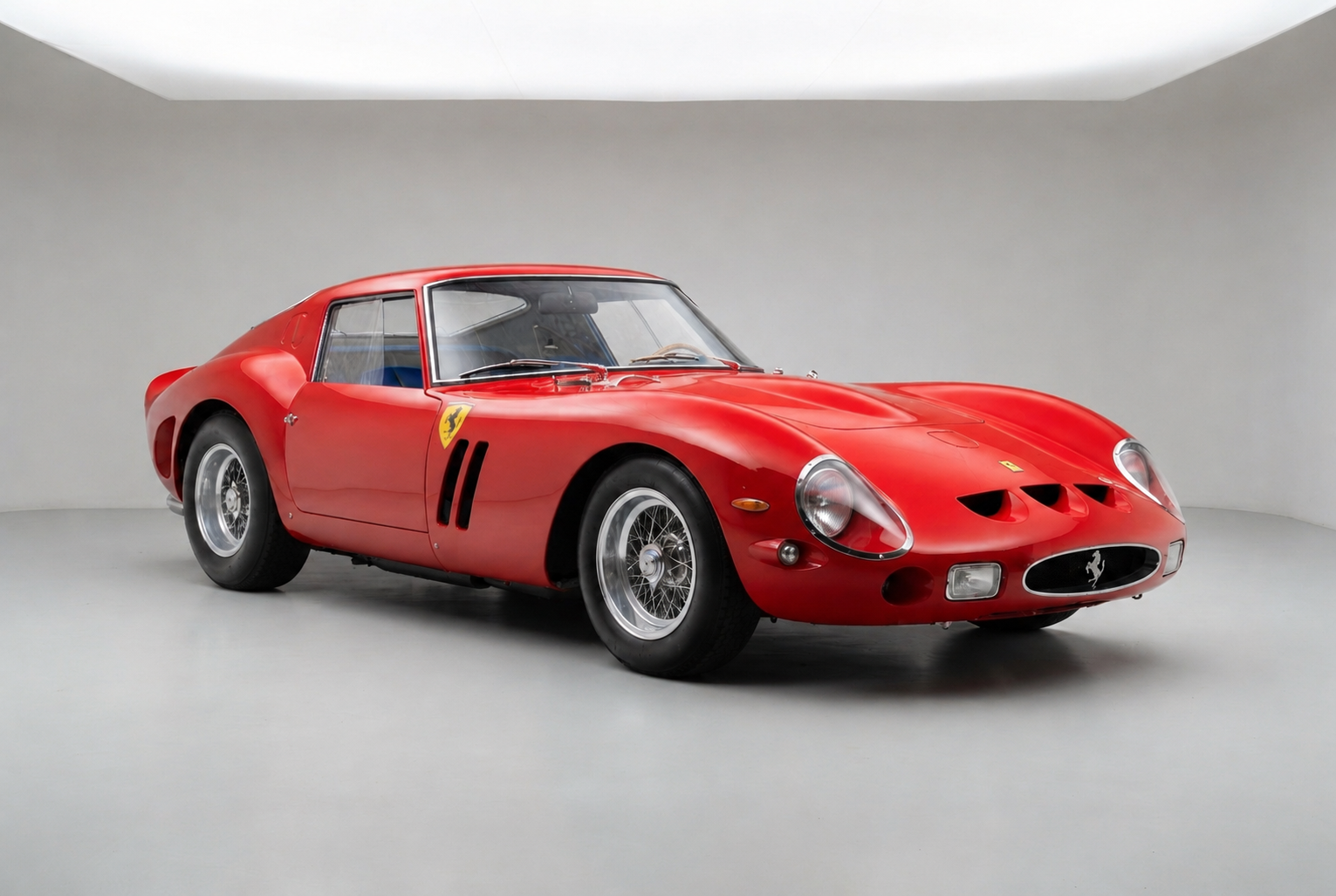 Red Ferrari 250 GTO Series I, front three-quarter studio view on a pale grey backdrop, showing the oval grille with prancing horse badge, covered headlamps, the three D-shaped nose panels, Borrani wire wheels and the yellow Scuderia shield on the door.