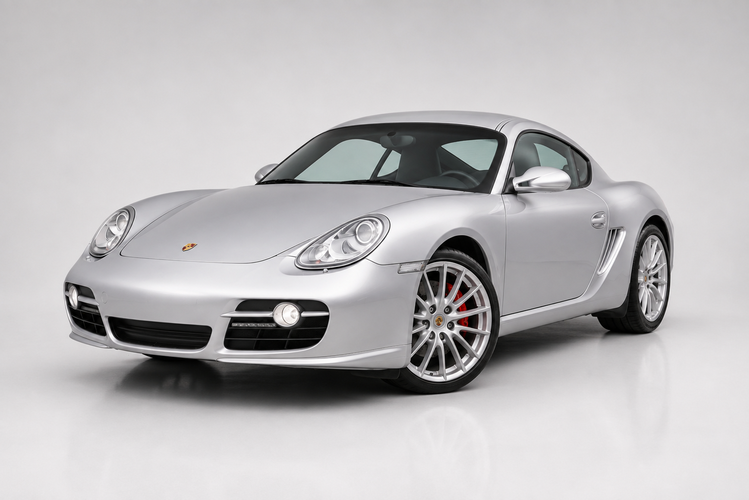 Black Porsche Cayman coupe in a studio setting, front three-quarter view showing the round headlights, low front bumper with LED daytime running strips, side air intake, black multi-spoke alloy wheels and red brake calipers.