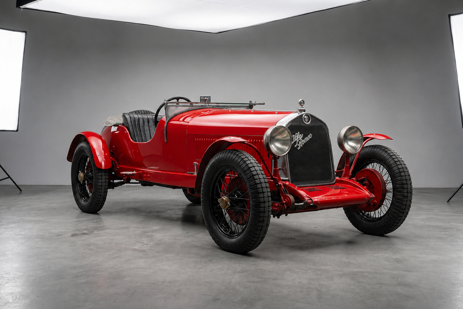 Red pre-war Alfa Romeo 6C open two-seater speedster in a photographic studio, front three-quarter view showing the tall chrome Alfa Romeo grille, twin polished headlamps, folded windscreen frame and black wire wheels.