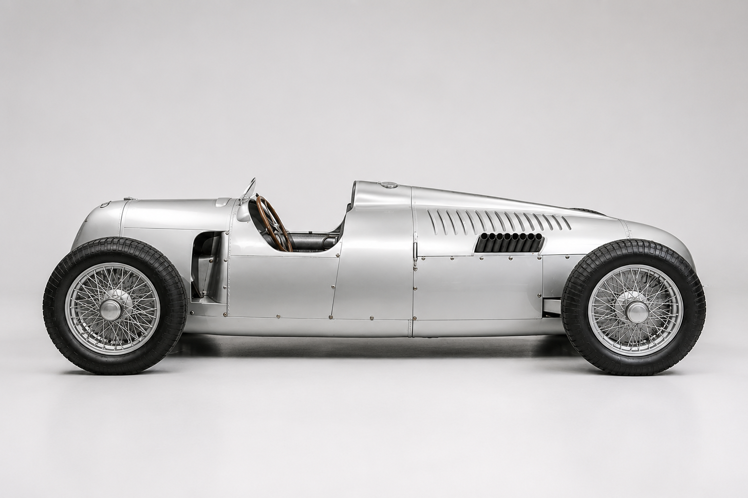 Silver Auto Union Type C grand prix car in side profile in a studio, showing the mid-engined streamlined body, louvred engine cover, exposed exhaust stubs and chrome wire wheels.