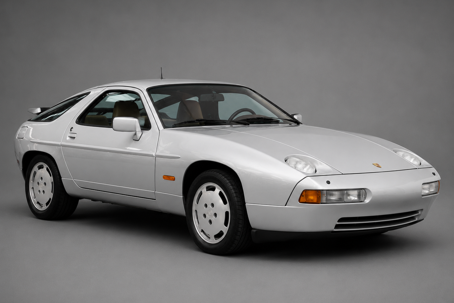 Studio three-quarter front view of a white Porsche 928 coupe with phone-dial alloy wheels and pop-up headlights raised, on a plain grey backdrop.