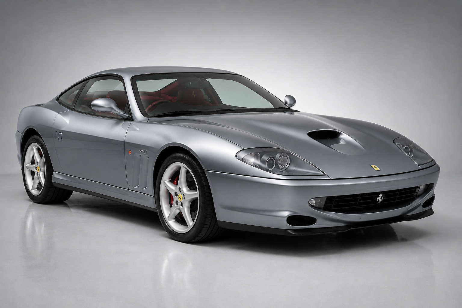 Silver Ferrari 550 Maranello coupe in a grey studio setting, front three-quarter view showing the long bonnet with central scoop, faired-in headlamps, oval lower grille, red brake calipers behind five-spoke alloy wheels and red leather interior.