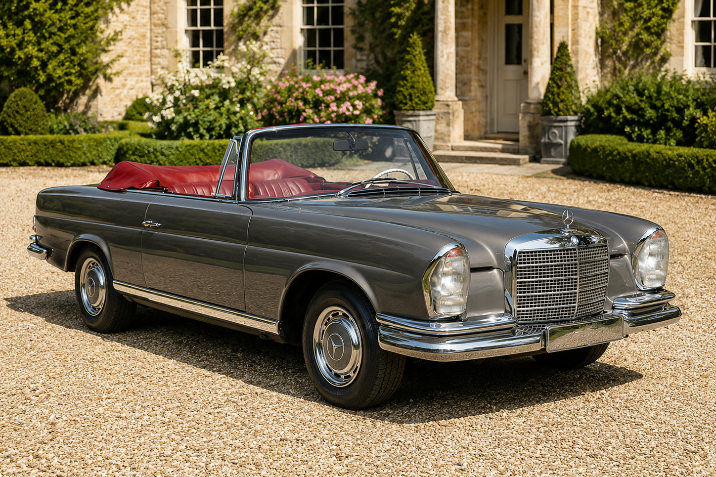 Mercedes-Benz 280 SE 3.5 Cabriolet (W111) in dark grey with red leather interior, front three-quarter view on a gravel driveway in front of a Cotswold-stone country house showing the final hand-built Stuttgart cabriolet in a concours / estate setting.