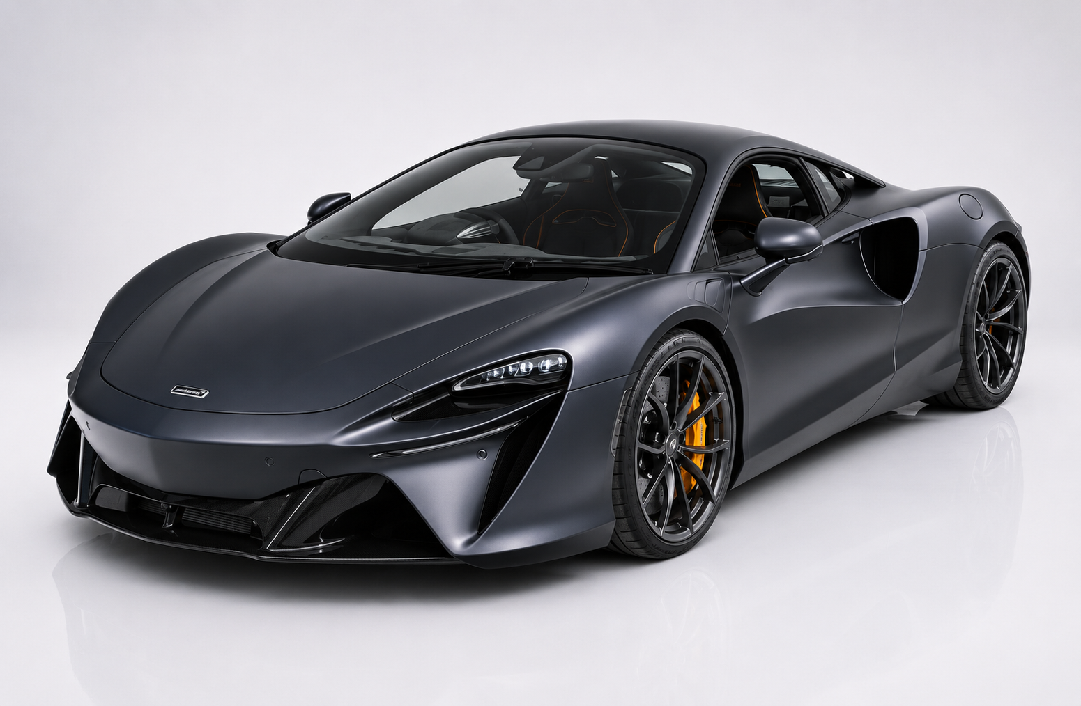 Dark matte-grey McLaren Artura in a studio setting, front three-quarter view showing the low mid-engine silhouette on the MCLA carbon platform, LED front lighting, prominent side air intake behind the door and dark multi-spoke wheels with yellow brake calipers — McLaren's first series-production V6 hybrid supercar.