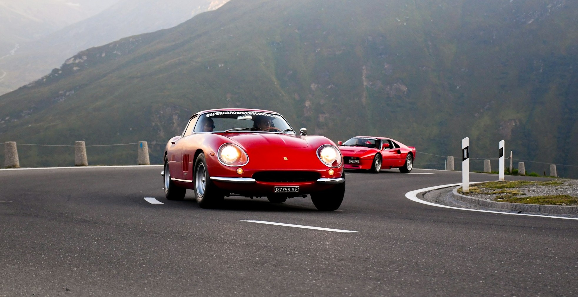 A red Ferrari 275 GTB and red Ferrari 288 GTO on a winding mountain road with green alpine peaks behind.
