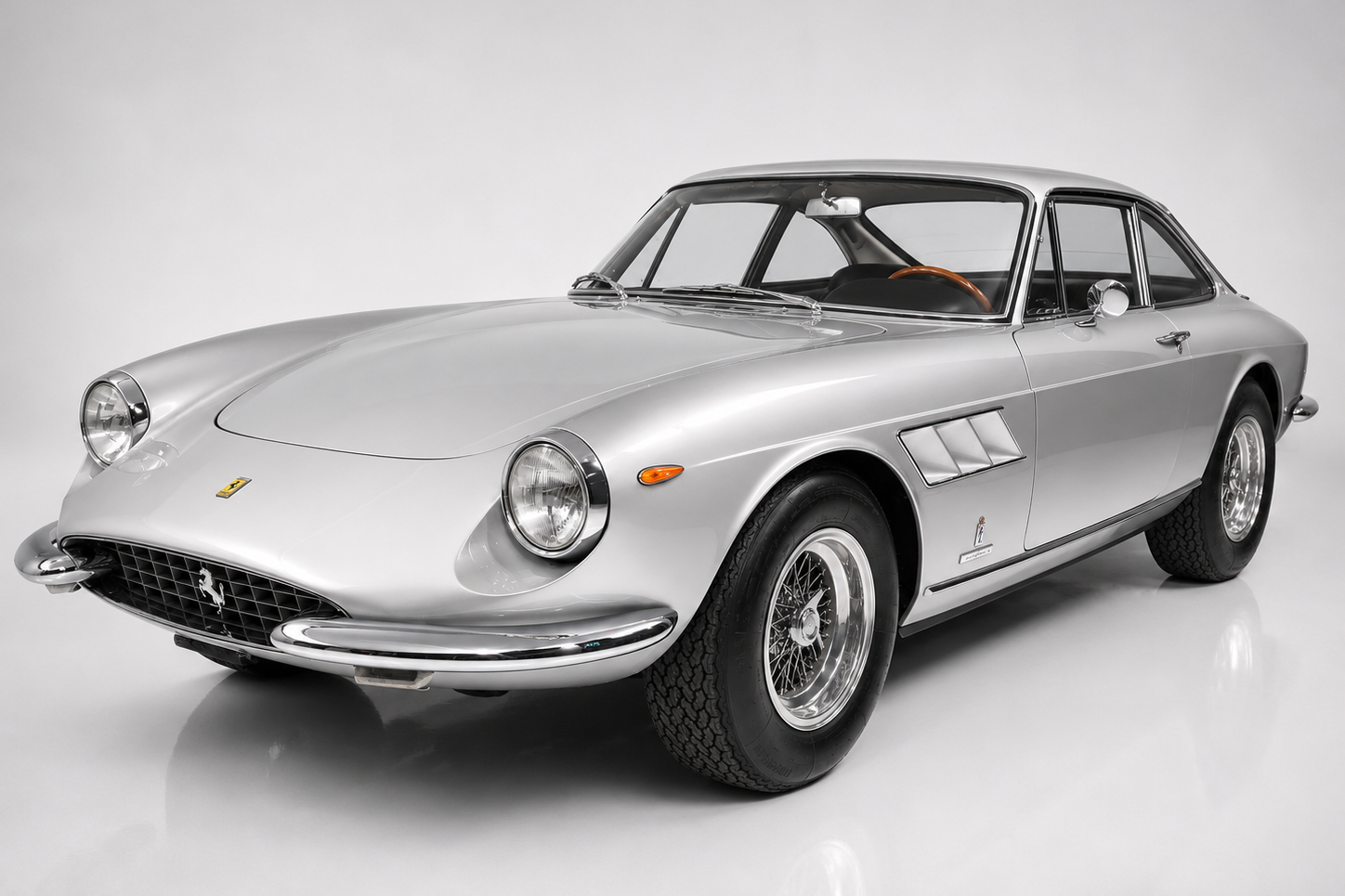 Silver Ferrari 330 GTC Pininfarina berlinetta, front three-quarter studio view showing the low nose with oval mesh grille and prancing horse badge, round headlamps, side fender vents, chrome bumperettes and chrome wire wheels on a plain pale backdrop.