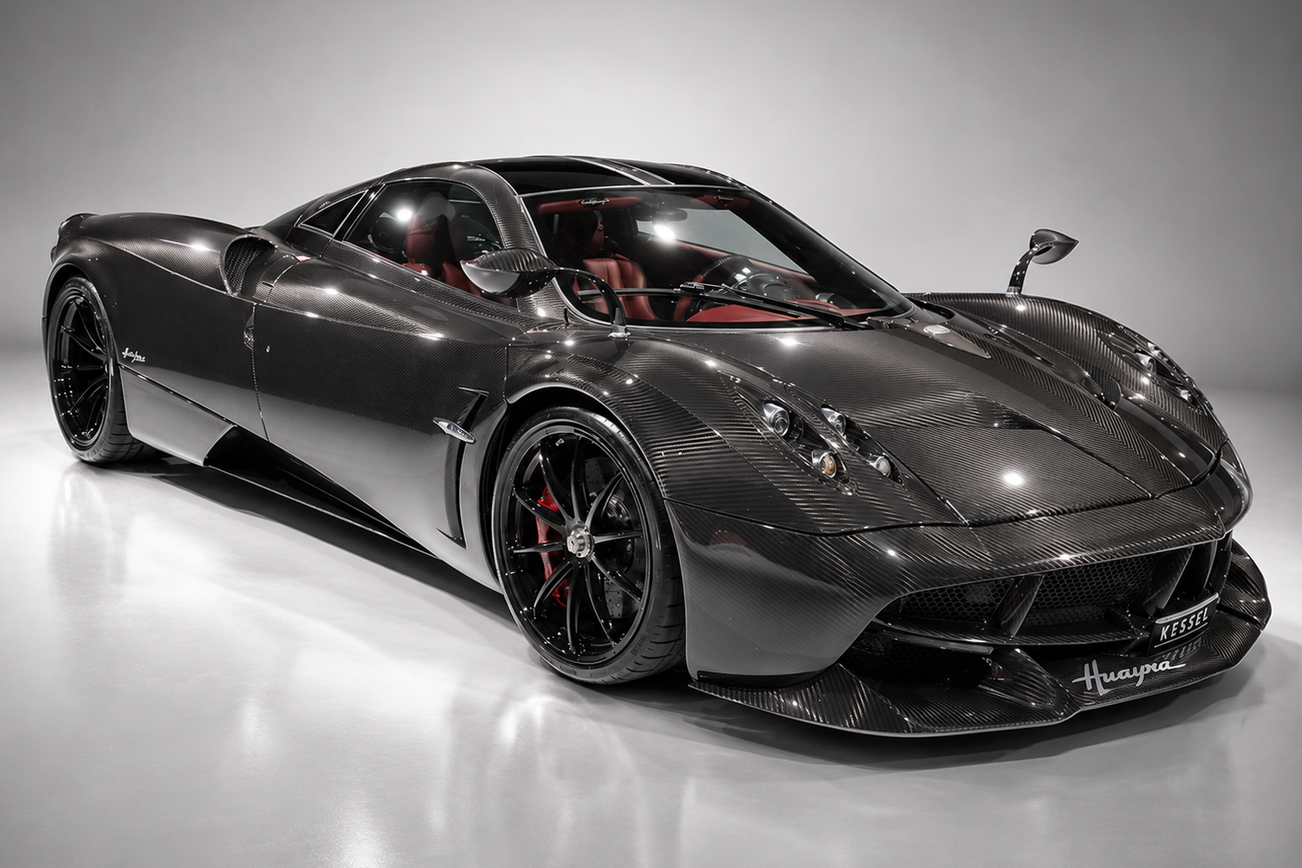Exposed carbon-fibre Pagani Huayra in a studio, front three-quarter view showing the active aerodynamic flaps, quad-headlamp clusters, sculpted side intakes, black multi-spoke wheels with red brake calipers and red leather interior visible through the glass canopy.