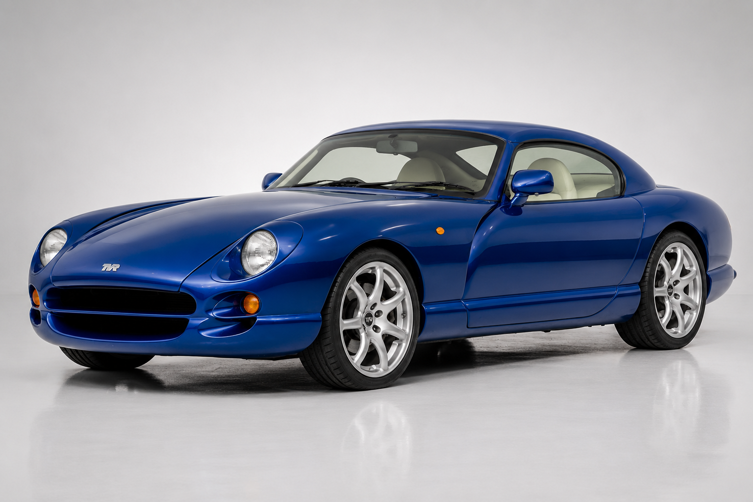 Blue TVR Cerbera in a studio setting, front three-quarter view showing the exposed round headlights, TVR badge on the smooth nose, sculpted composite bodywork and multi-spoke silver alloy wheels.
