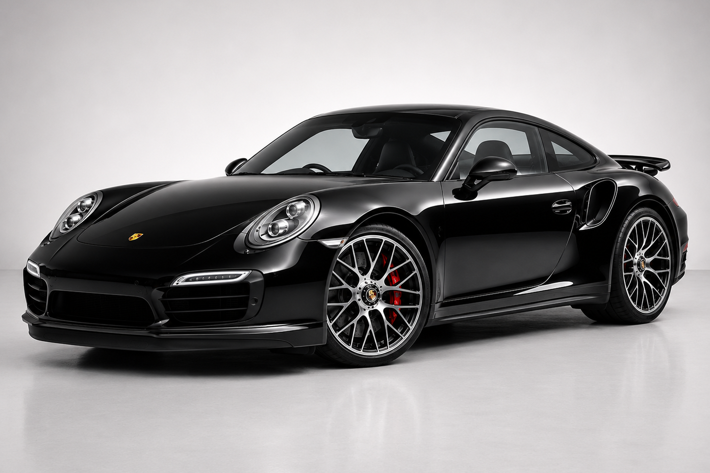 Black Porsche 911 (991) Turbo coupe in a studio setting, front three-quarter view showing the round headlights with LED daytime running strips, low front bumper with side intakes, fixed rear wing and multi-spoke alloy wheels with red brake calipers.