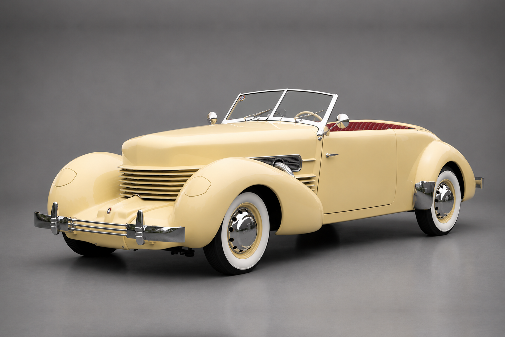 Cream Cord 810/812 Sportsman cabriolet with top down and red interior, front three-quarter studio view on a plain grey backdrop showing the coffin-nose bonnet, wraparound louvred grille, chrome bumper and whitewall tyres.