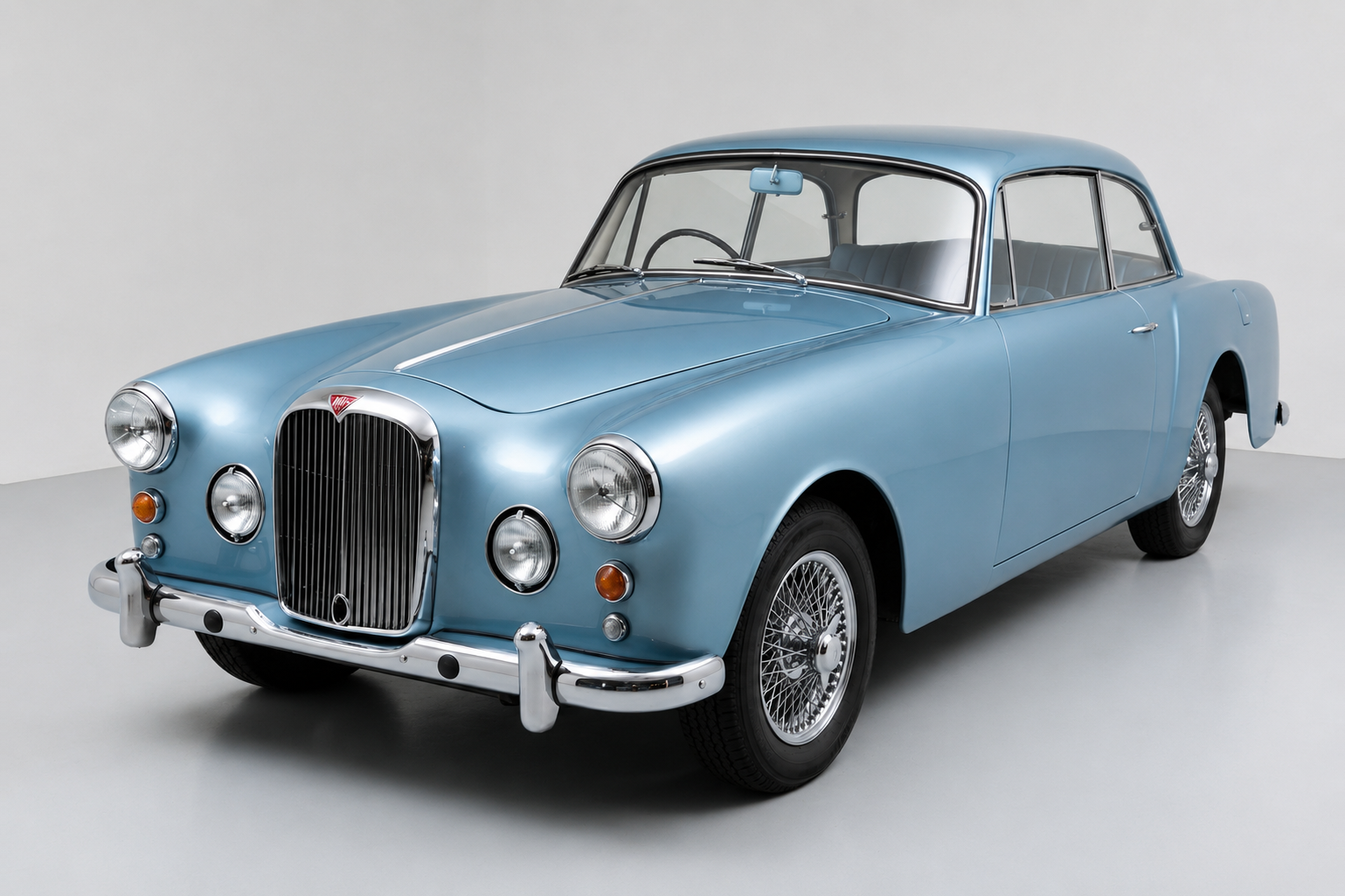 Light metallic blue Alvis TD21 two-door saloon in a studio, front three-quarter view showing the upright chrome grille with red Alvis badge, paired chrome headlamps and auxiliary lamps, chrome bumper with overriders and centre-lock wire wheels.