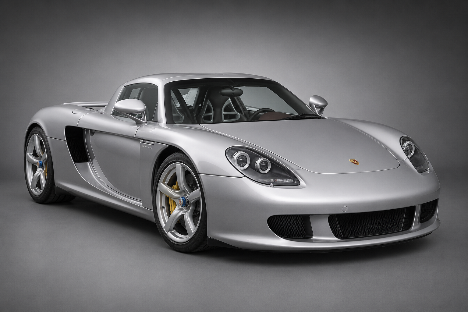Silver Porsche Carrera GT in a grey studio setting, front three-quarter view showing the removable targa roof panel, yellow ceramic brake calipers and five-spoke alloy wheels.