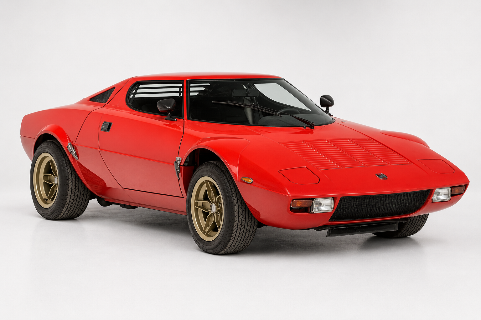 Red Lancia Stratos HF coupe in a bright studio setting, front three-quarter view showing the short wedge body, wraparound windscreen, slatted rear window louvres, black grille with twin round headlights and gold five-spoke alloy wheels.