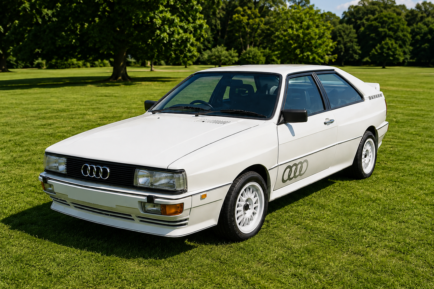 White Audi Ur-Quattro Turbo coupe on a manicured lawn, front three-quarter view showing the four-ring grille, twin rectangular headlamps, front spoiler, white lattice alloy wheels and side 'Audi rings' decal.