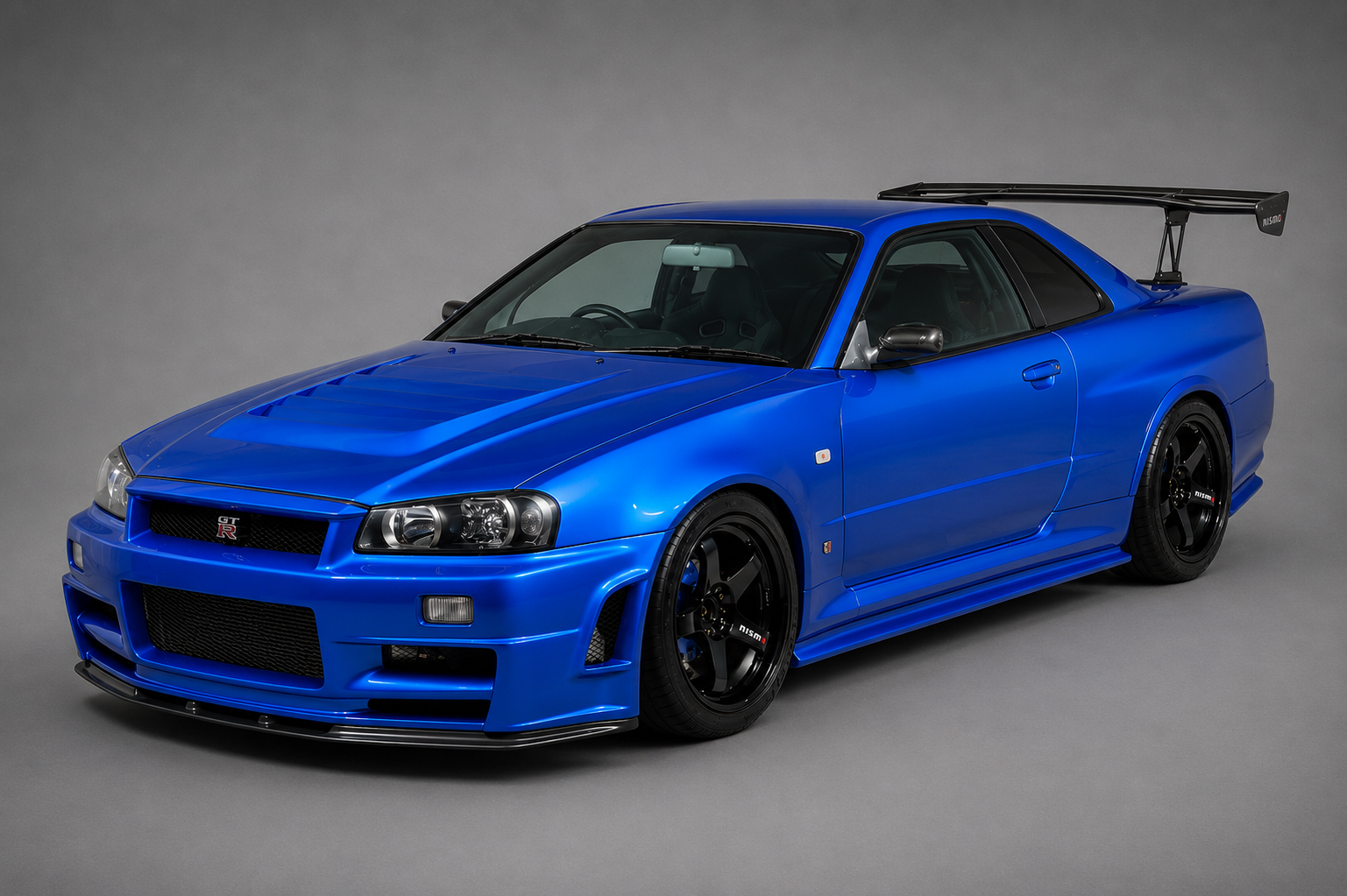 Studio three-quarter front view of a Bayside Blue Nissan Skyline GT-R R34 coupe with black multi-spoke alloy wheels and a large rear wing, on a plain grey backdrop.