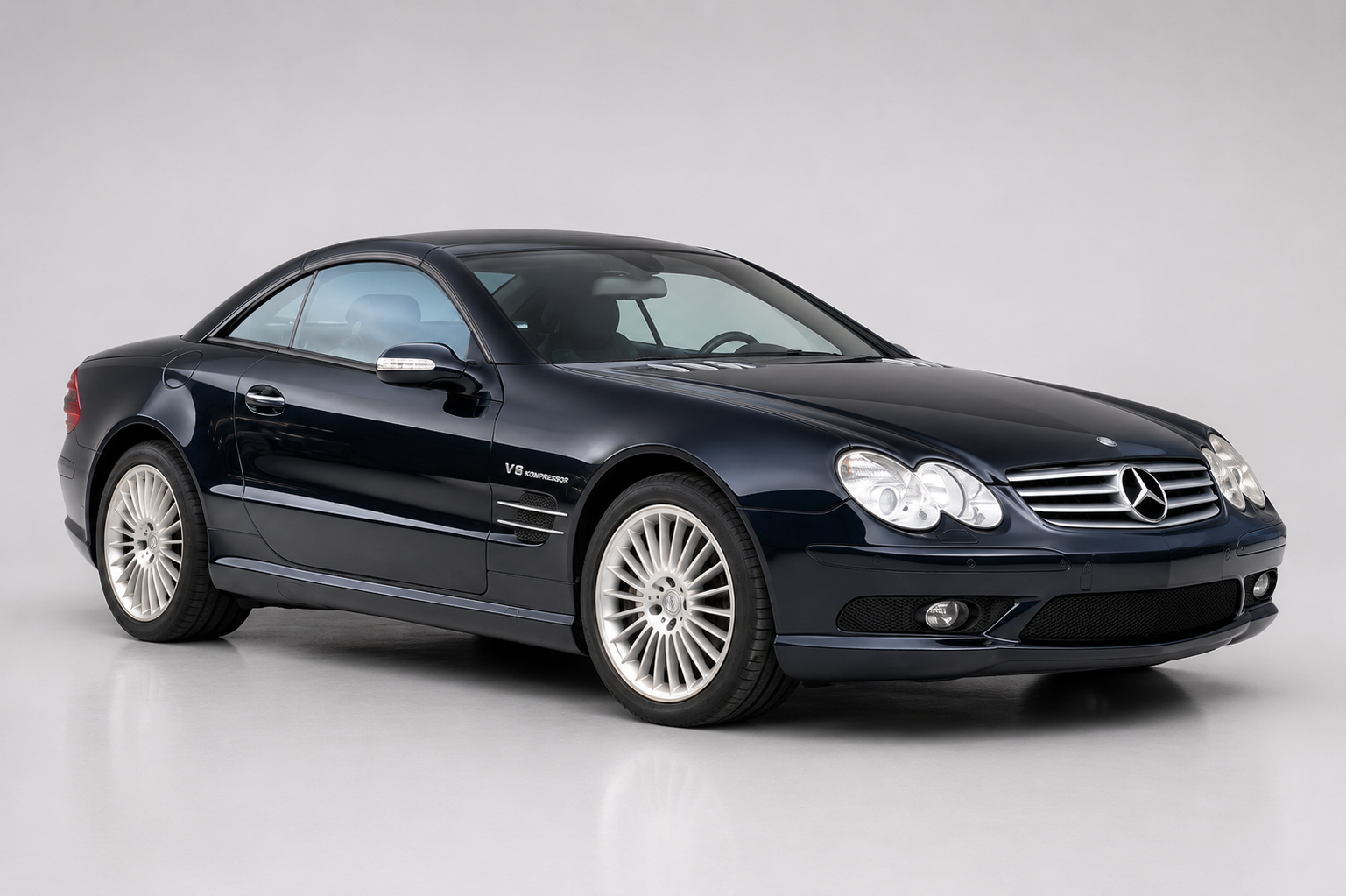 Silver Mercedes-Benz SL 63 AMG (R230) with hardtop raised in a plain studio setting, front three-quarter view showing the AMG front apron with LED daytime running lights, bonnet vents, V8 badging on the front wing and multi-spoke AMG alloy wheels.