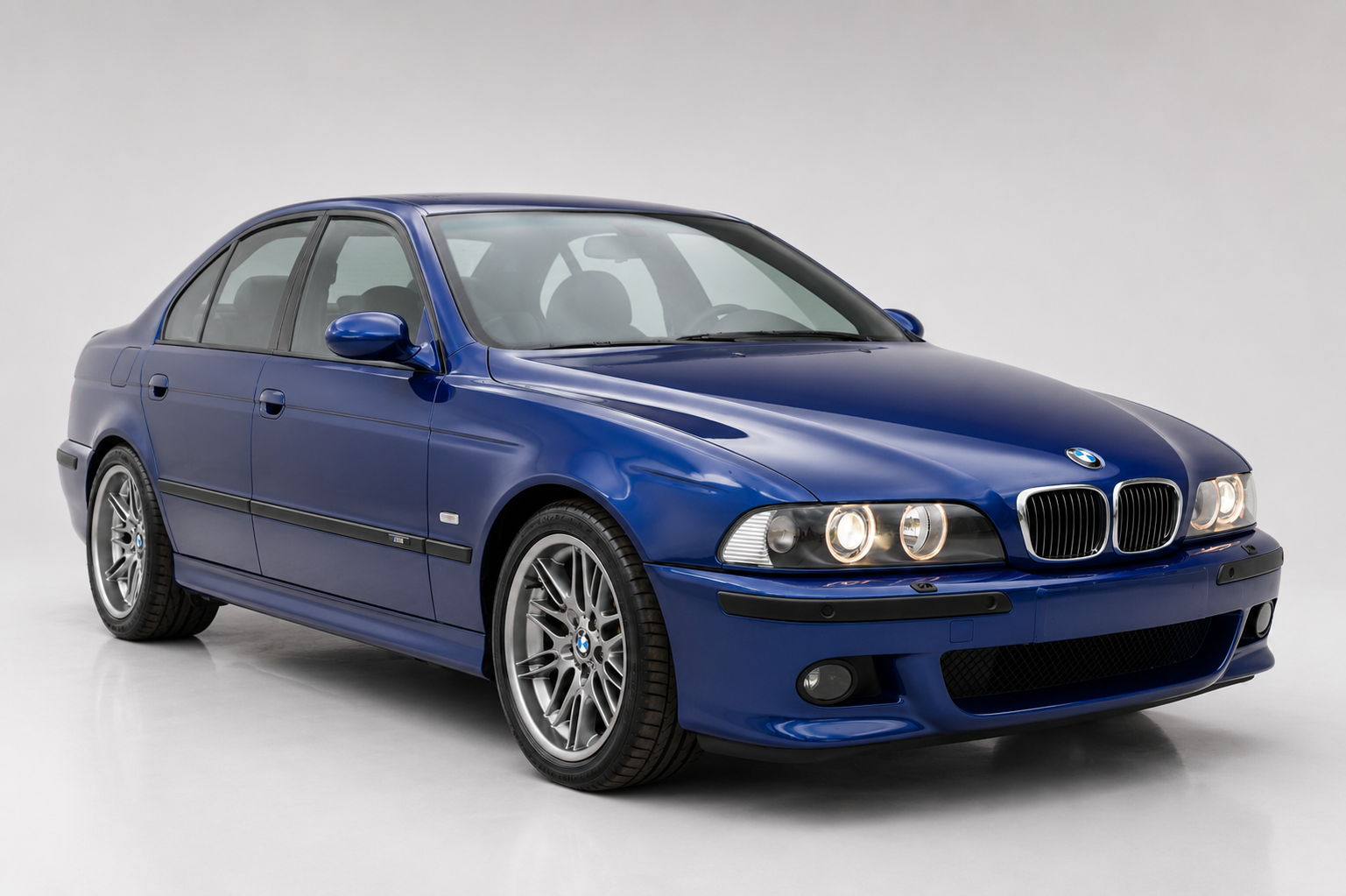 Blue BMW M5 (E39) saloon in a grey studio setting, front three-quarter view showing the twin-kidney grille, quad round headlamps, side gill vent, multi-spoke alloy wheels and the muscular executive-saloon proportions.