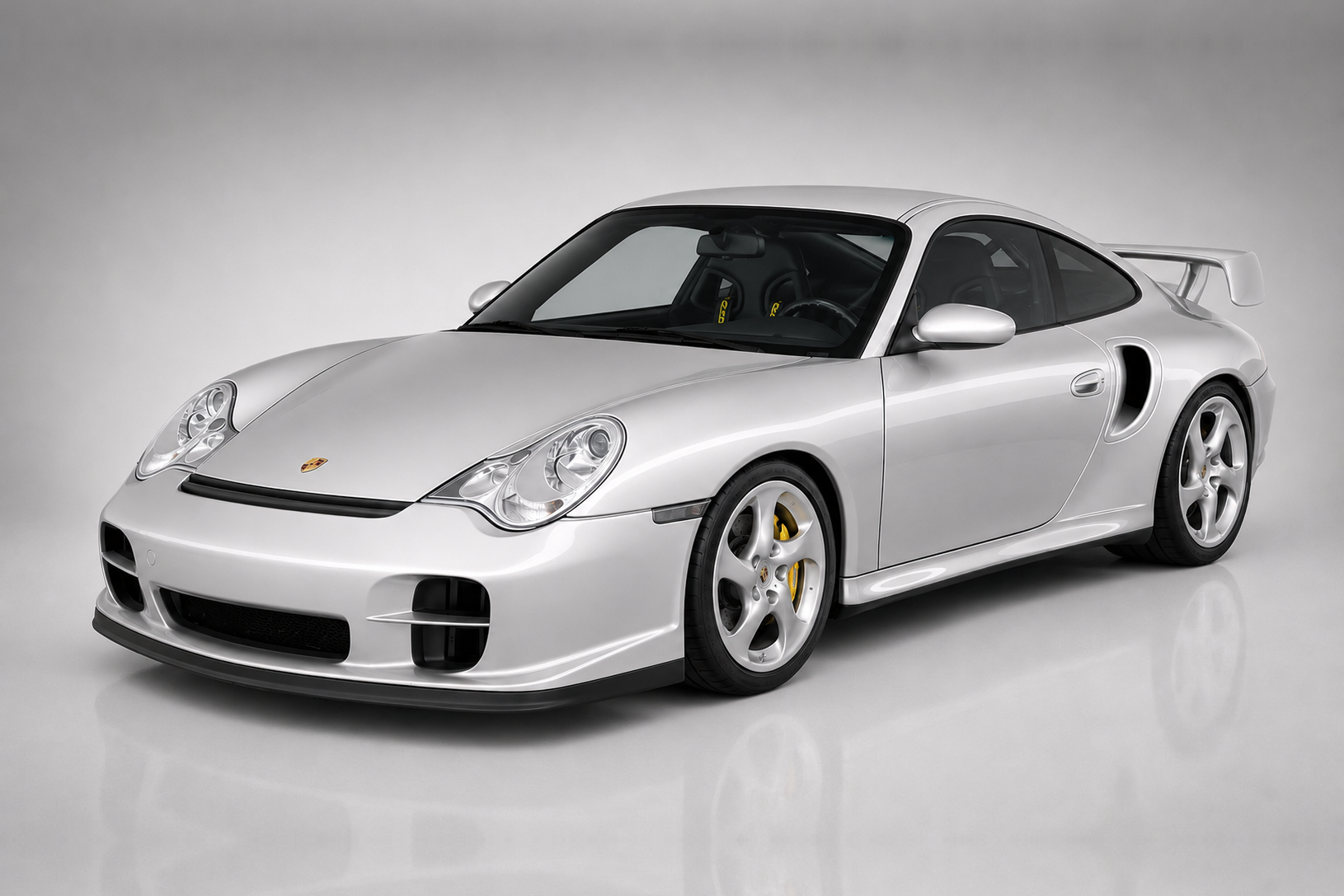 Silver Porsche 911 GT2 (996) in a studio, front three-quarter view showing the deep front bumper with brake cooling ducts, side intercooler intakes, fixed rear wing and multi-spoke silver wheels.
