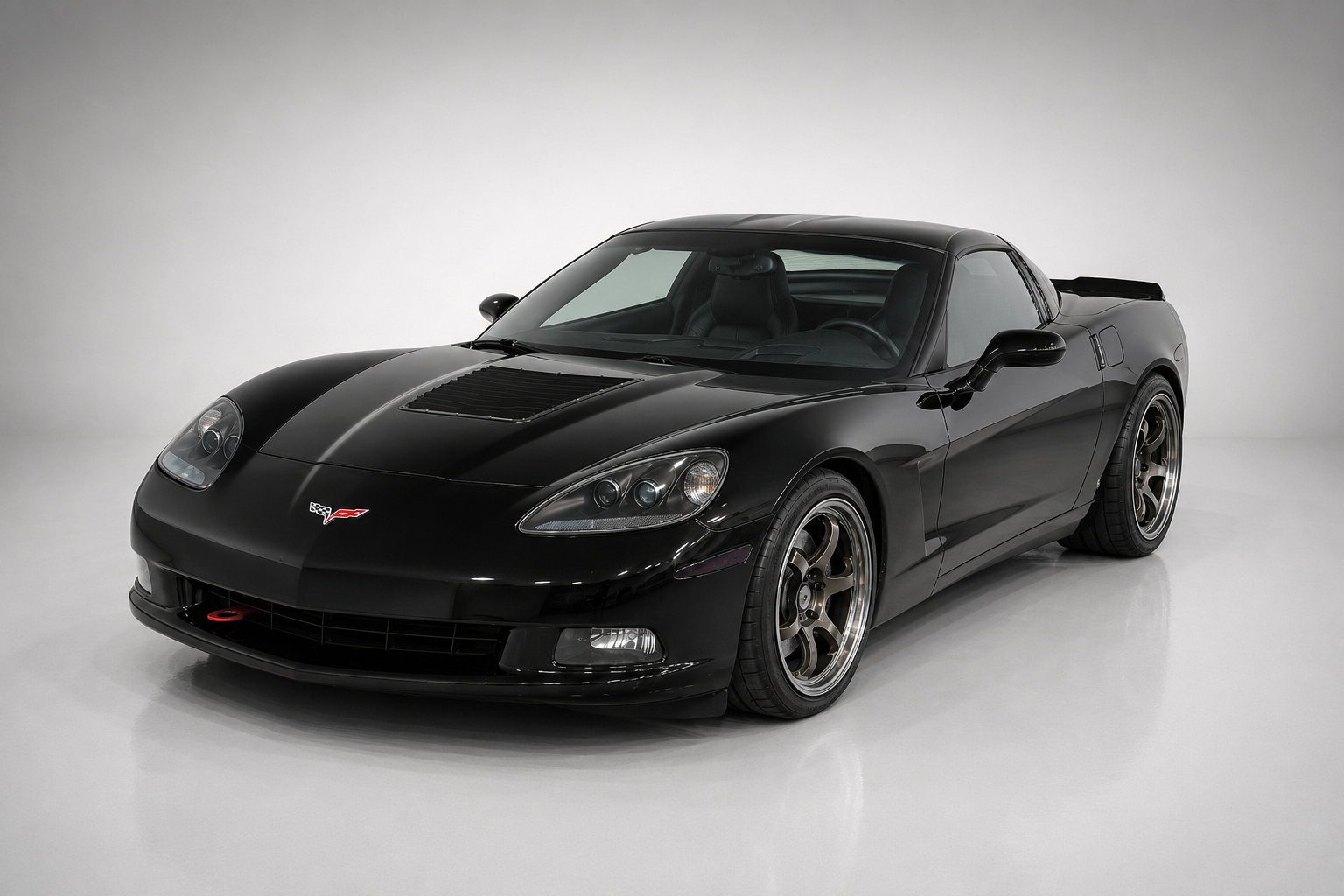 Black Chevrolet Corvette C6 coupe in a studio setting, front three-quarter view, showing exposed projector headlights (first modern Corvette without pop-ups), bonnet with intake and multi-spoke bronze alloy wheels.
