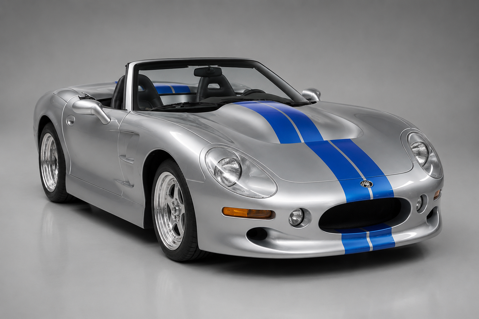 Silver Shelby Series 1 roadster with blue centre stripes, front three-quarter studio view showing the low aluminium body, round quad headlights, oval grille aperture and polished multi-spoke alloy wheels.