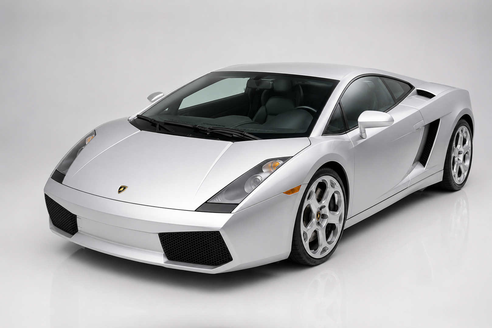 Silver Lamborghini Gallardo coupe in a bright studio, front three-quarter view showing the sharp early-Gallardo bodywork, rounded headlamp clusters, wide low nose with black lower intakes, side air scoop and silver alloy wheels.