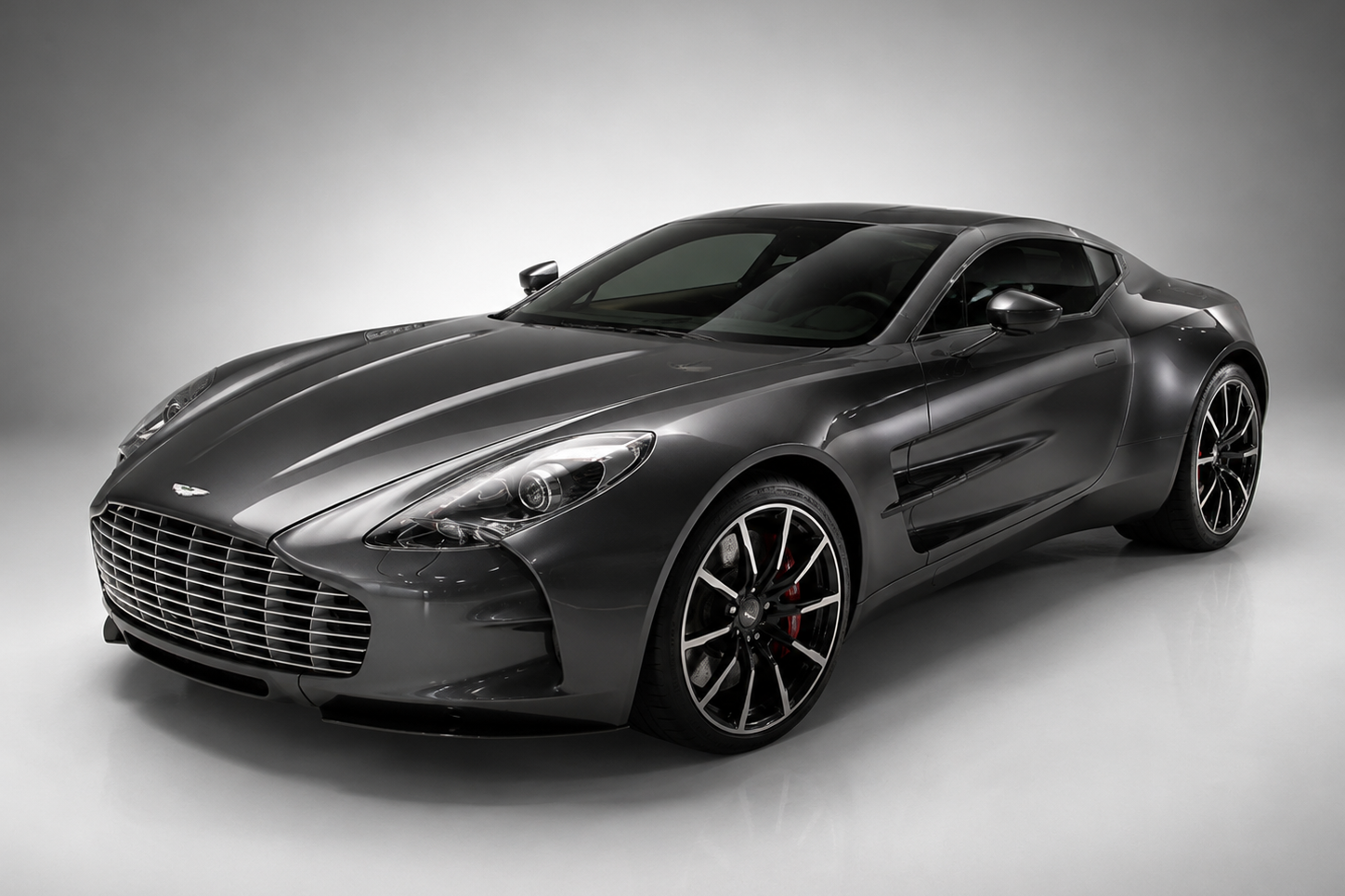 Grey Aston Martin One-77 in a studio setting, front three-quarter view showing the sculpted aluminium bodywork, wide chrome grille, distinctive LED headlamps and black multi-spoke alloy wheels with red brake calipers.