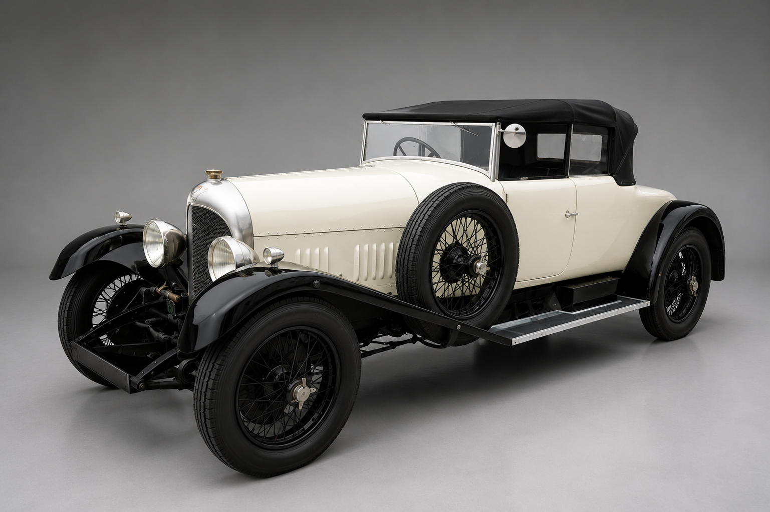 Cream and black 1920s Bentley 3 Litre with drophead coupe coachwork and black canvas hood raised, front three-quarter studio view showing the tall radiator, side-mounted spare wheel, wire wheels and black wings.