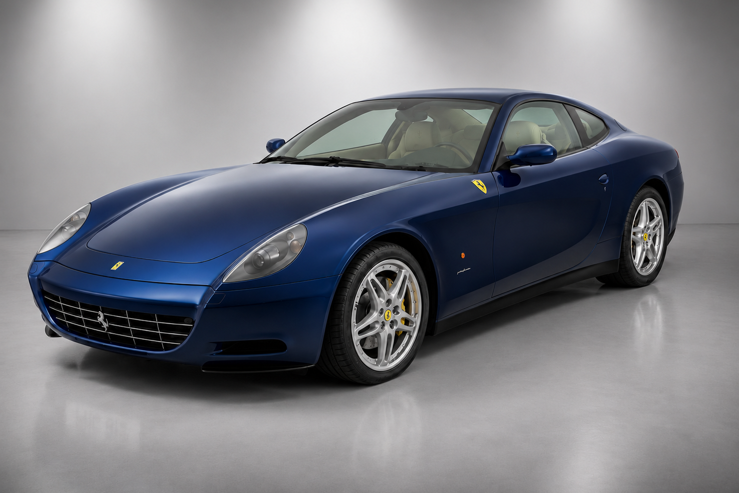 Dark blue Ferrari 612 Scaglietti coupe in a grey studio setting, front three-quarter view showing the Pininfarina four-seat V12 GT bodywork, oval mesh grille, cream leather interior and silver multi-spoke alloy wheels.
