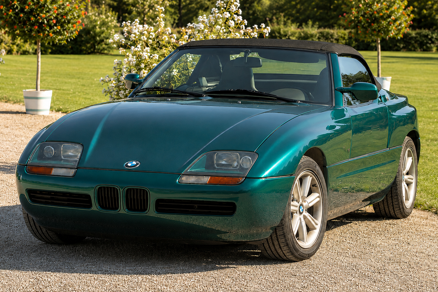 Dark green BMW Z1 with the black soft top raised, parked on gravel in a garden setting, front three-quarter view showing the BMW kidney grille, compact headlamps, black lower intake and the unusual drop-door roadster silhouette.