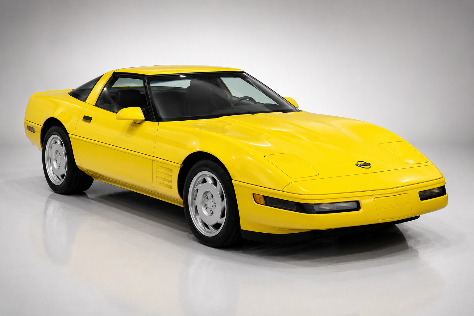 Yellow Chevrolet Corvette C4 coupe in a light studio setting, front three-quarter view, showing pop-up headlights raised, body-colour bumpers and silver five-spoke sawblade alloy wheels.