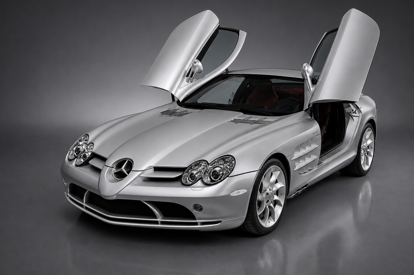 Silver Mercedes-Benz SLR McLaren coupe in a studio setting, both butterfly doors raised, showing the long bonnet with side exhaust vents, quad projector headlamps, chrome three-pointed-star grille and red leather interior.
