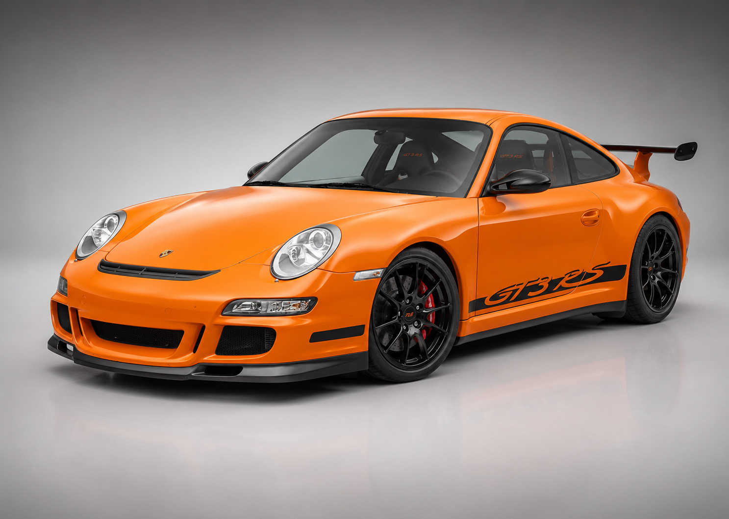 Orange Porsche 911 GT3 RS (997) in a grey studio, front three-quarter view showing the black GT3 RS side script, gloss-black centre-lock wheels and adjustable carbon rear wing.
