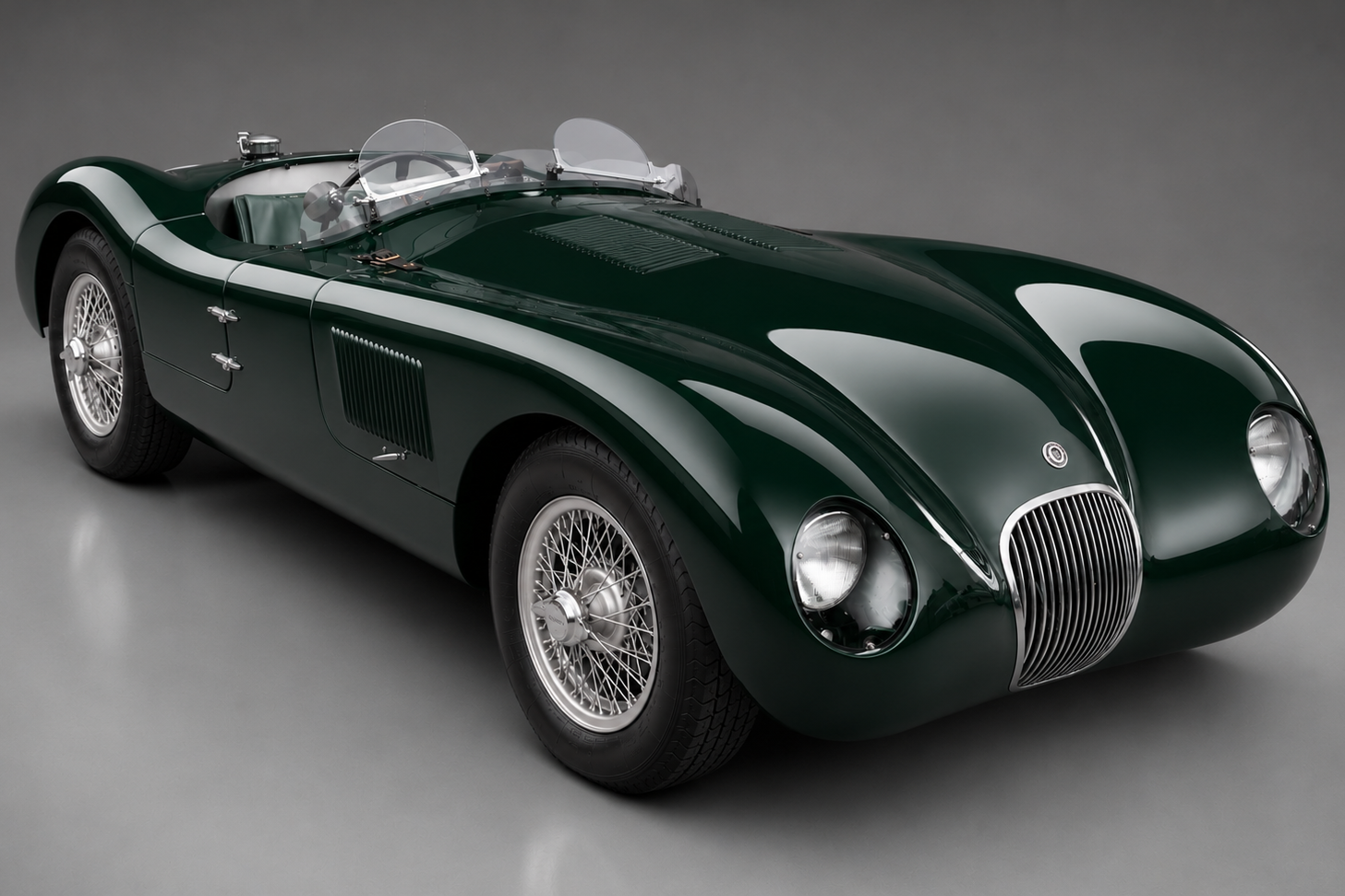 Jaguar C-Type in British Racing Green, front three-quarter studio view on a grey backdrop showing the aluminium body, wire wheels, twin aero-screens and chrome grille.
