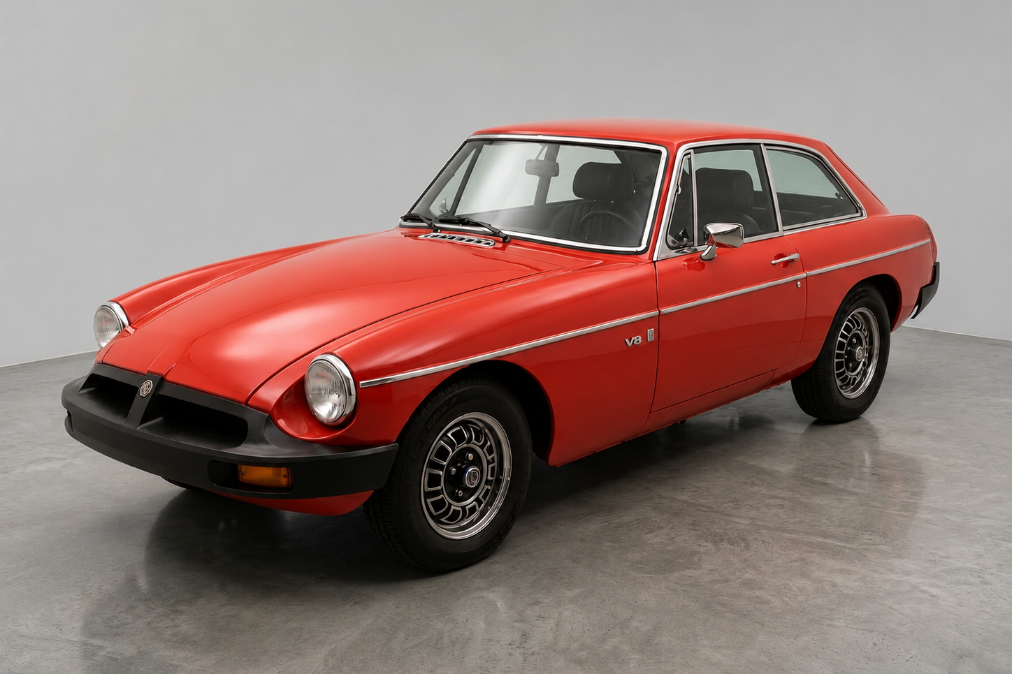 Red MGB GT V8 coupé with black front bumper, chrome side trim and a V8 badge on the front wing, front three-quarter view in a studio against a neutral background, showing the composite alloy wheels and chrome window surrounds.