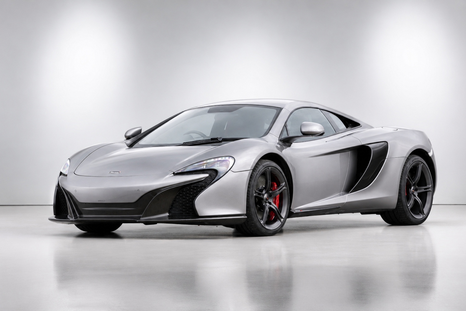 Silver McLaren 650S coupé in a plain studio setting, front three-quarter view showing the P1-derived front end, dihedral doors, side intake ducts, dark multi-spoke alloy wheels and red brake calipers.