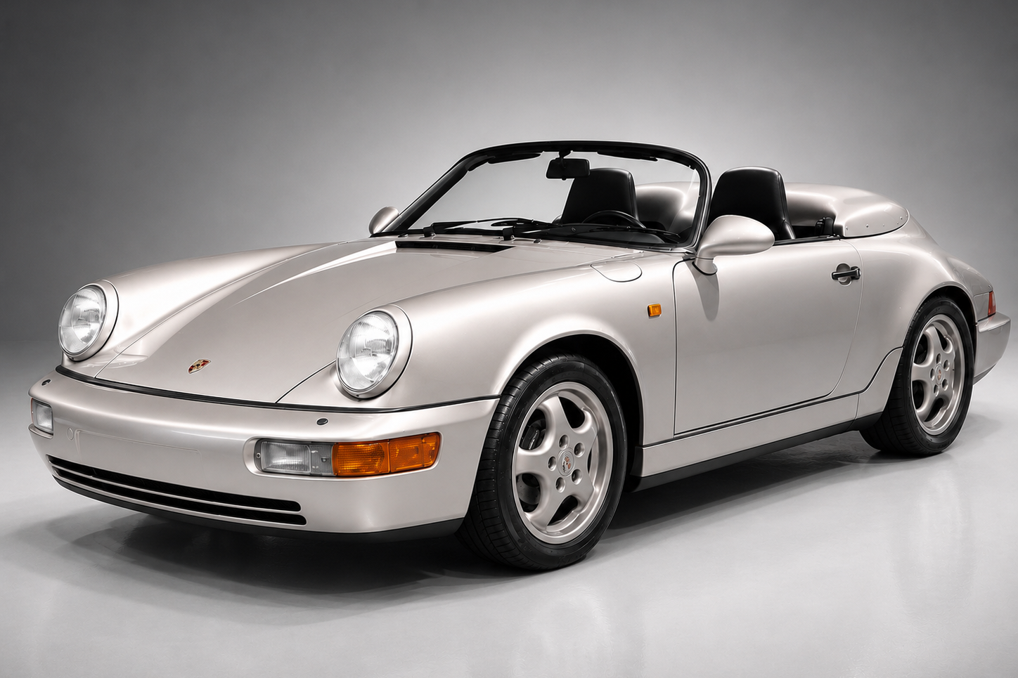 Silver Porsche 911 (964) Speedster in a studio setting, front three-quarter view showing the round headlights, integrated body-colour bumpers, low raked windscreen, black interior with roof stowed and five-spoke Cup-style alloy wheels.