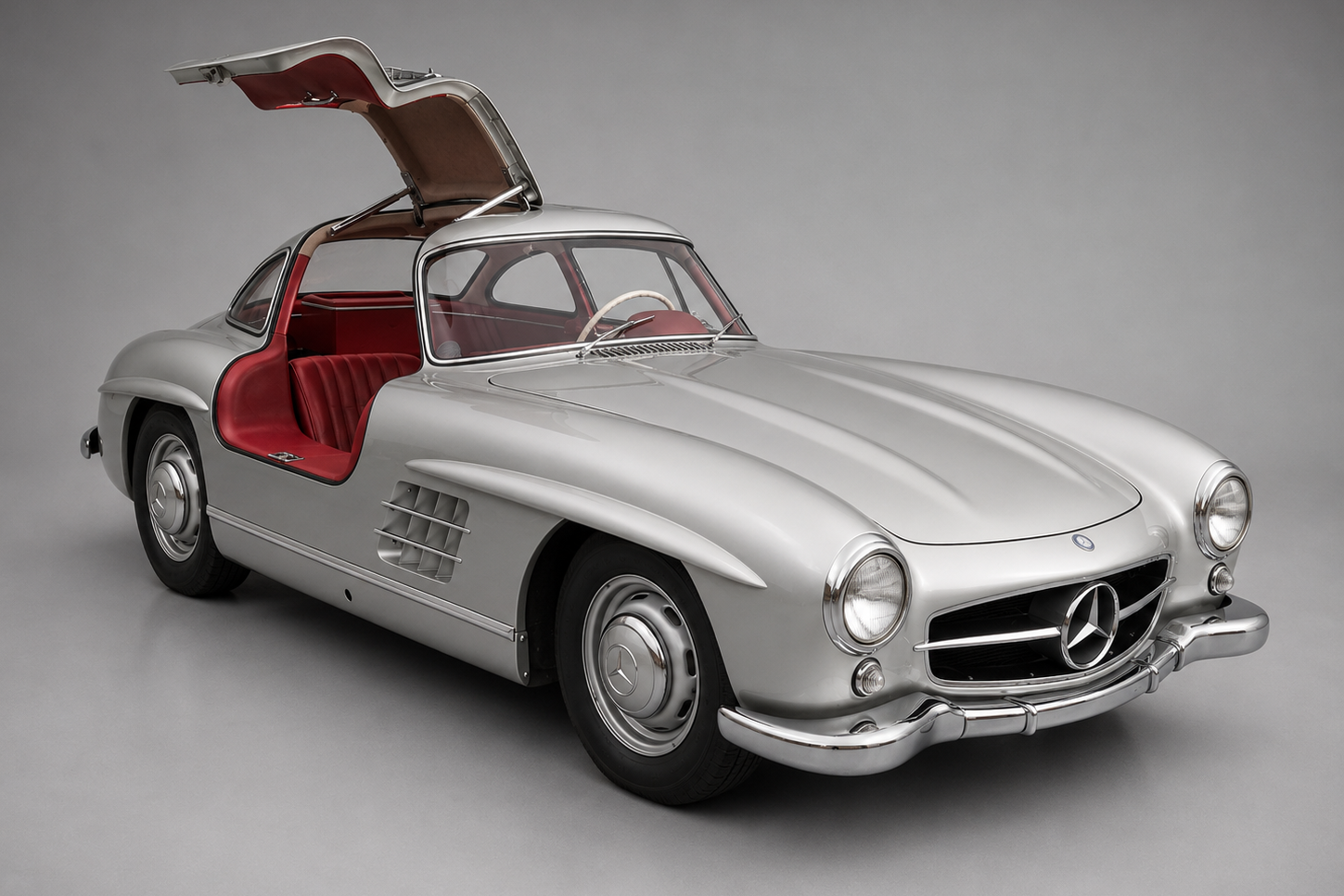 Silver Mercedes-Benz 300 SL Gullwing with one upward-opening door raised, showing the red interior, photographed against a neutral grey studio backdrop.